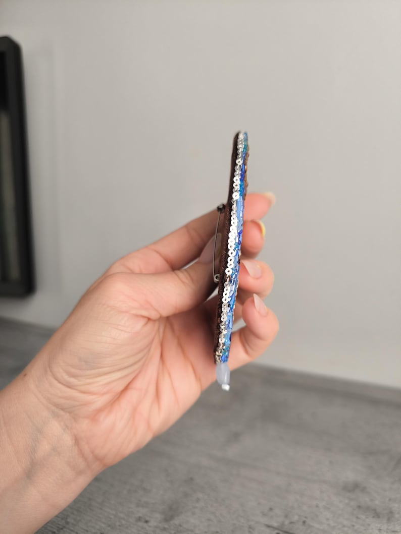 May include: A handmade brooch with a dark brown base, adorned with blue and silver sequins. The brooch has a pin on the back for easy attachment. The design features a gradient of blue sequins, creating a shimmering effect.