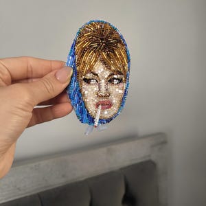 May include: A beaded brooch featuring a portrait of a woman with blonde hair and a blue headscarf, smoking a cigarette. The face is detailed with small beads in various colors, creating a mosaic effect. The brooch is oval-shaped.
