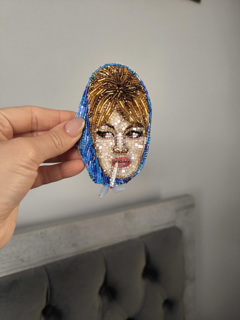 May include: A beaded brooch featuring a portrait of a woman with blonde hair and a blue headscarf, smoking a cigarette. The face is detailed with small beads in various colors, creating a mosaic effect. The brooch is oval-shaped.