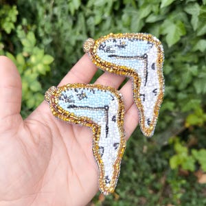 May include: Two beaded brooches shaped like melting clocks, featuring light blue faces, black clock hands, and white and gold beaded details. The brooches are reminiscent of a surrealist art style.
