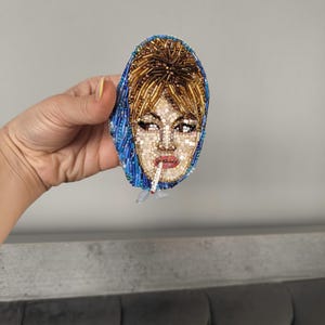 May include: A beaded brooch depicting a woman with blonde hair and a blue headscarf, smoking a cigarette. The brooch is oval-shaped and features intricate beadwork in various colors, including gold, blue, and flesh tones. The woman's face is detailed with beads.
