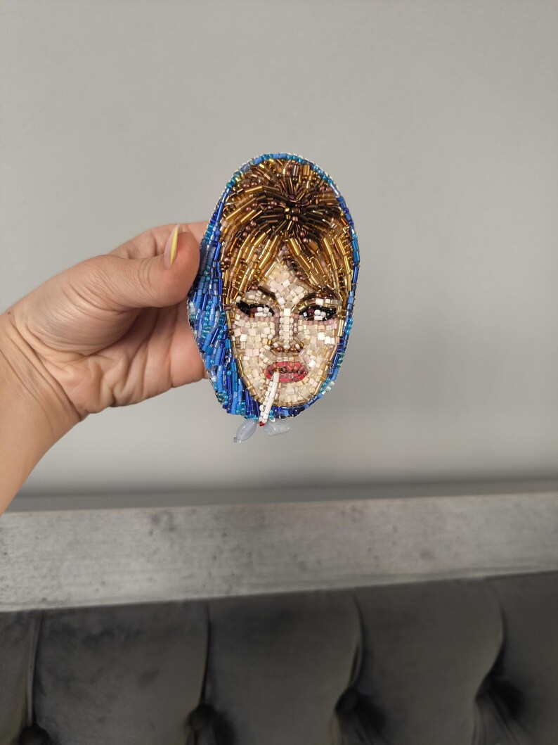 May include: A beaded brooch depicting a woman with blonde hair and a blue headscarf, smoking a cigarette. The brooch is oval-shaped and features intricate beadwork in various colors, including gold, blue, and flesh tones. The woman's face is detailed with beads.