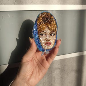 May include: Oval-shaped beaded brooch featuring a portrait of a woman with blonde hair and a blue headscarf. The face is detailed with small beads, and the brooch has a light blue and gold color scheme. The woman is smoking a cigarette.