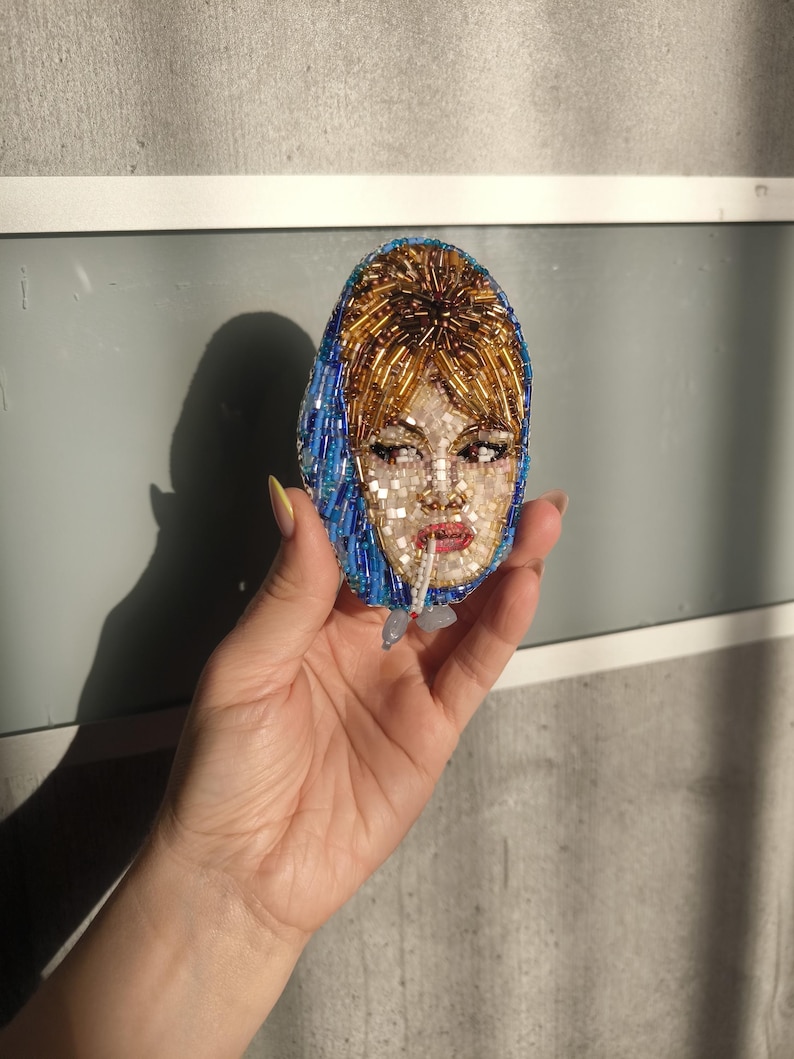 May include: Oval-shaped beaded brooch featuring a portrait of a woman with blonde hair and a blue headscarf. The face is detailed with small beads, and the brooch has a light blue and gold color scheme. The woman is smoking a cigarette.