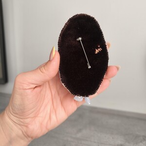 May include: A dark brown velvet brooch with a silver pin backing. The brooch is oval-shaped and has a white trim. Small, light-colored beads are attached to the bottom of the brooch.