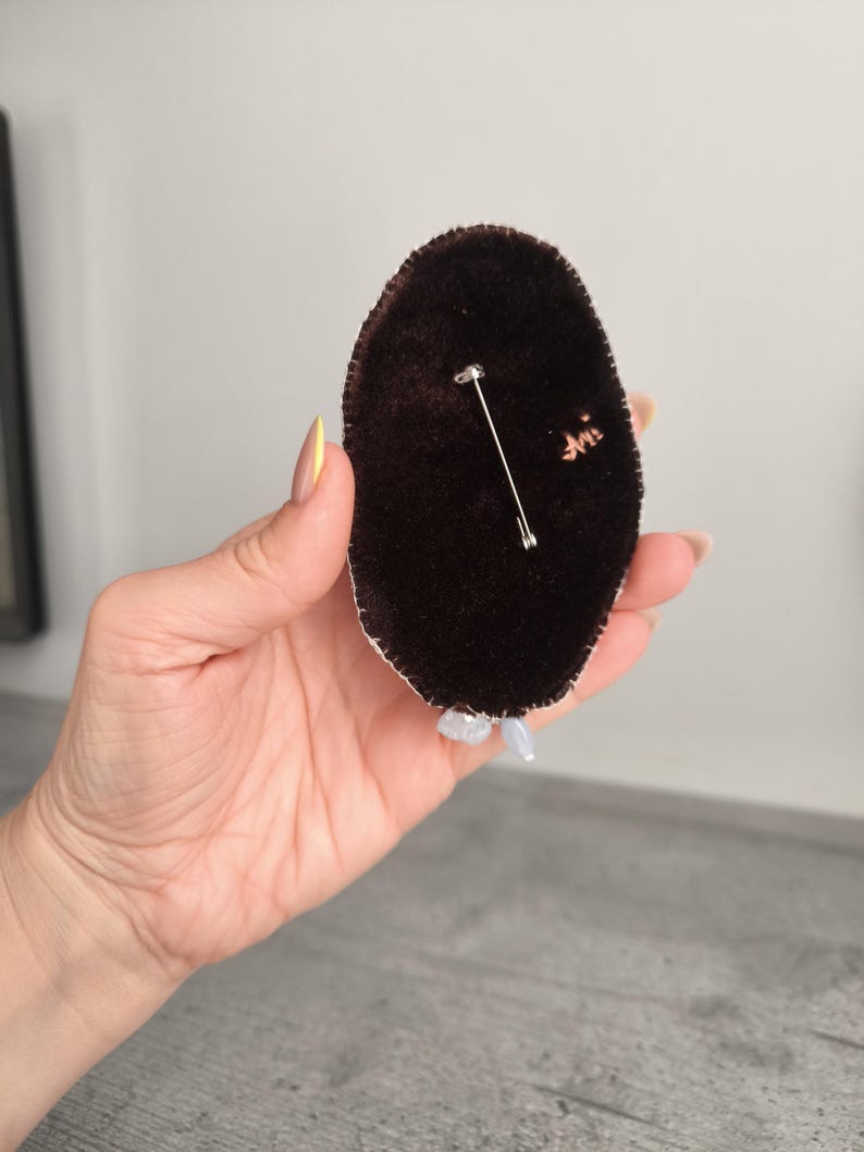 May include: A dark brown velvet brooch with a silver pin backing. The brooch is oval-shaped and has a white trim. Small, light-colored beads are attached to the bottom of the brooch.