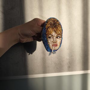 May include: A beaded brooch featuring a portrait of a woman with red hair and a blue headscarf. The face is intricately detailed with beads, and the brooch has a vintage aesthetic. The woman is smoking a cigarette.