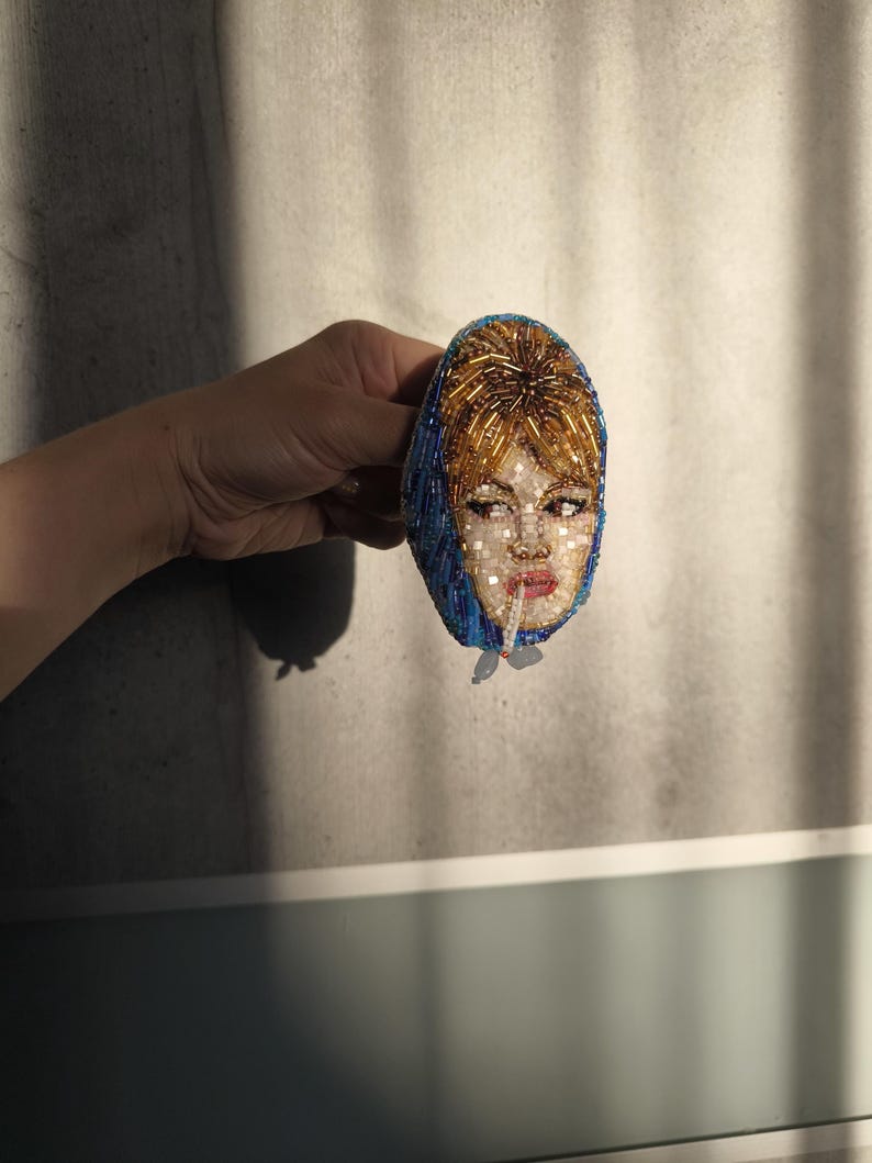 May include: A beaded brooch featuring a portrait of a woman with red hair and a blue headscarf. The face is intricately detailed with beads, and the brooch has a vintage aesthetic. The woman is smoking a cigarette.