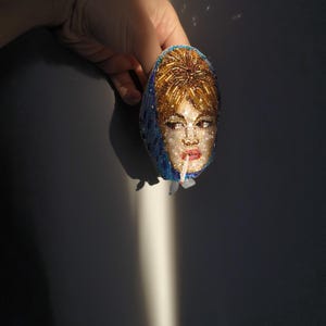May include: An oval-shaped brooch featuring a beaded portrait of a woman with blonde hair and red lipstick. The brooch has a blue headscarf and is held by a hand against a dark background. The portrait is made with small, colorful beads.