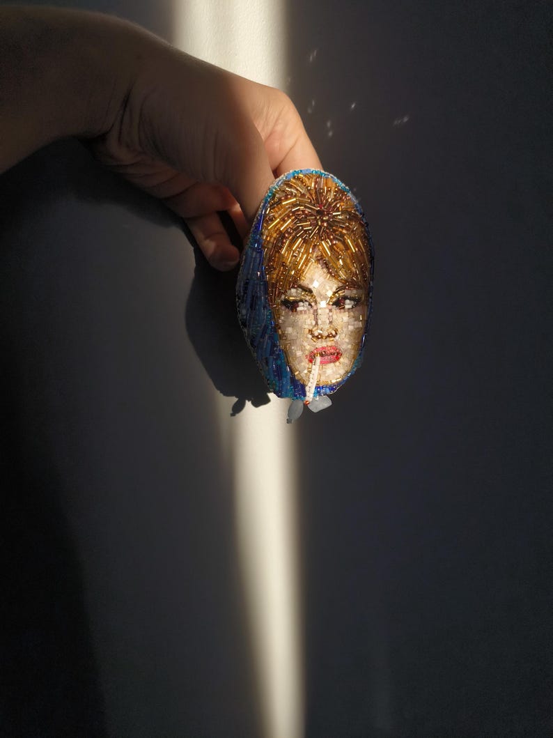 May include: An oval-shaped brooch featuring a beaded portrait of a woman with blonde hair and red lipstick. The brooch has a blue headscarf and is held by a hand against a dark background. The portrait is made with small, colorful beads.