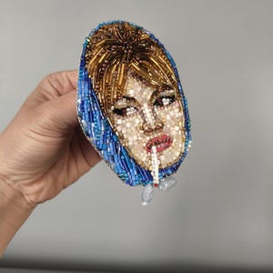 May include: A beaded brooch featuring a portrait of a woman with blonde hair and a blue headscarf, smoking a cigarette. The brooch is oval-shaped and made with various colored beads, including gold, blue, and white.