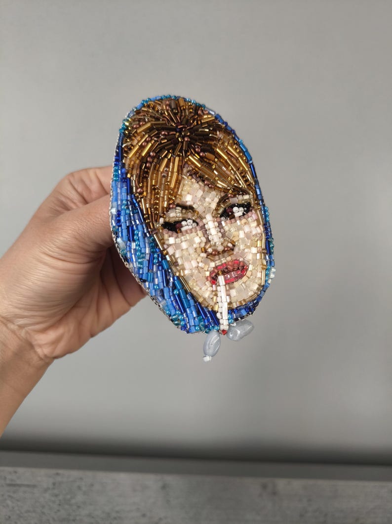 May include: A beaded brooch featuring a portrait of a woman with blonde hair and a blue headscarf, smoking a cigarette. The brooch is oval-shaped and made with various colored beads, including gold, blue, and white.