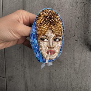 May include: Oval-shaped beaded brooch featuring a woman's face with a cigarette. The face is crafted with white and beige beads, red lips, and brown eyes. The hair is golden, and the head covering is blue. The brooch is likely handmade.