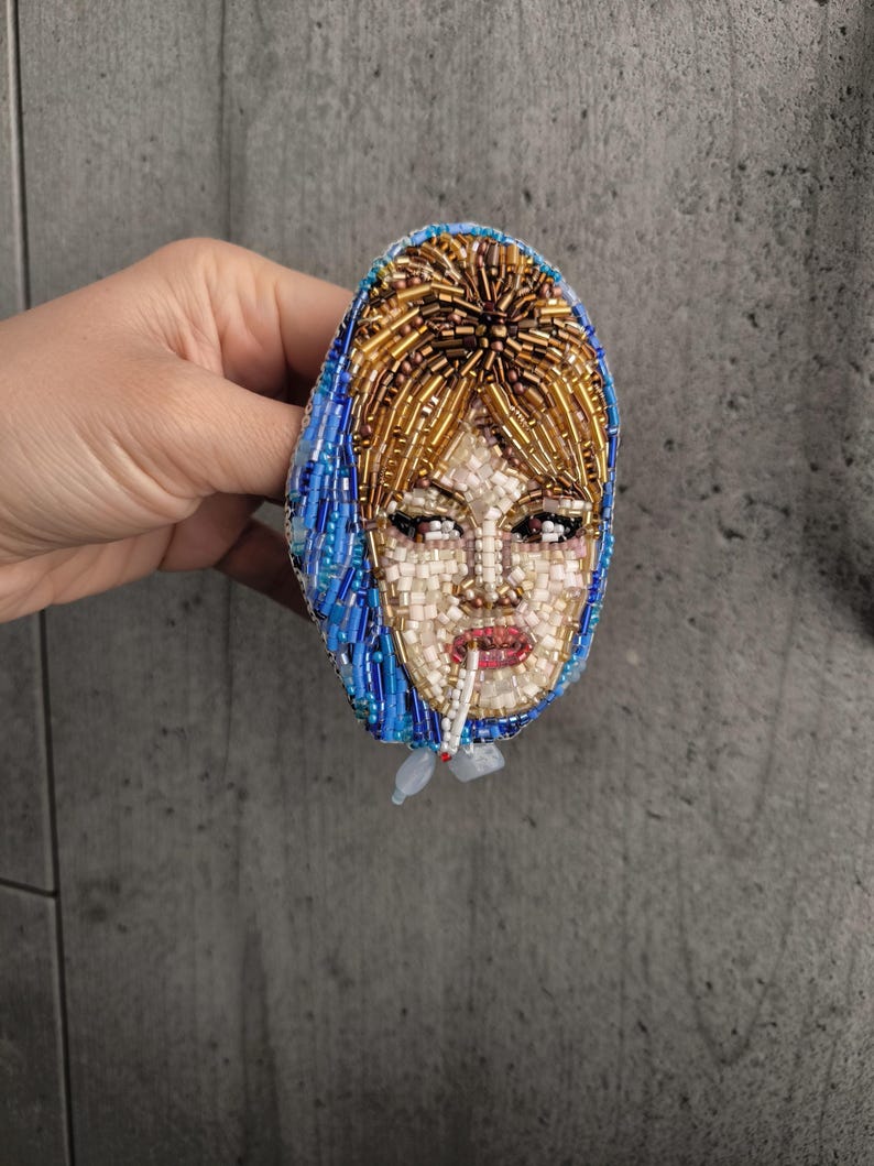 May include: Oval-shaped beaded brooch featuring a woman's face with a cigarette. The face is crafted with white and beige beads, red lips, and brown eyes. The hair is golden, and the head covering is blue. The brooch is likely handmade.