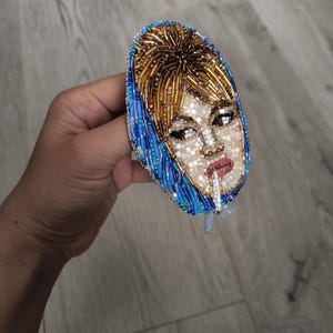 May include: A beaded brooch featuring a portrait of a woman with blonde hair and a blue headscarf, smoking a cigarette. The brooch is oval-shaped and made with various colored beads, including gold, blue, and white.