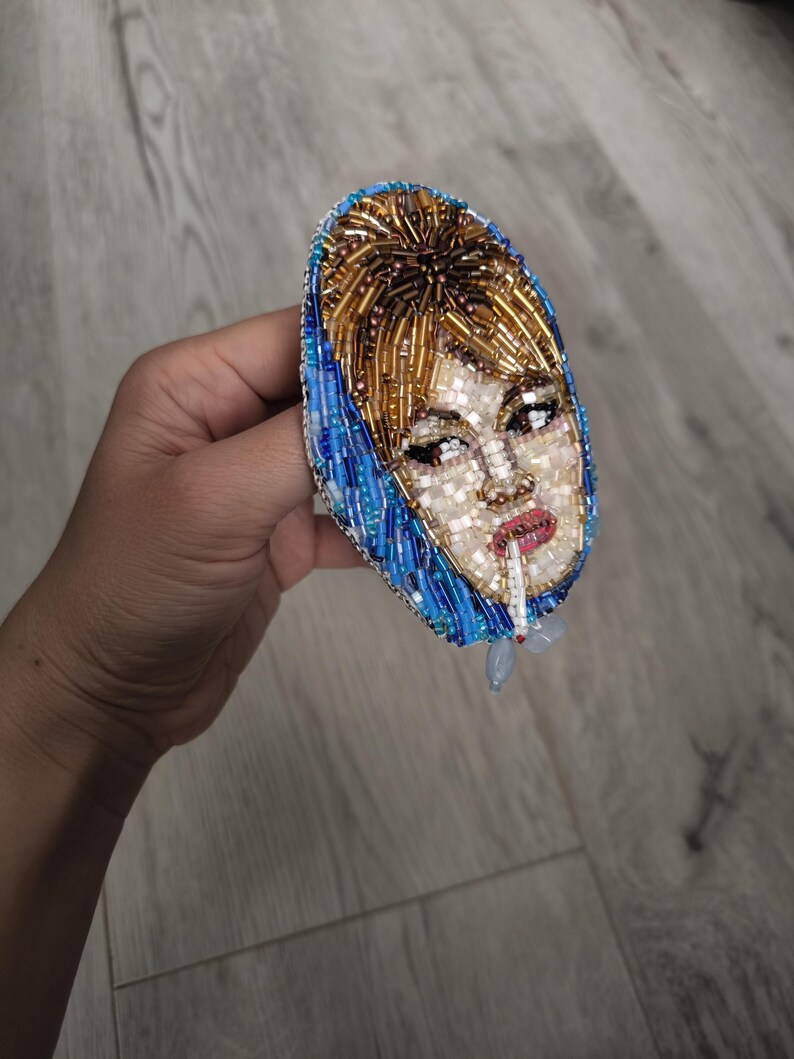 May include: A beaded brooch featuring a portrait of a woman with blonde hair and a blue headscarf, smoking a cigarette. The brooch is oval-shaped and made with various colored beads, including gold, blue, and white.