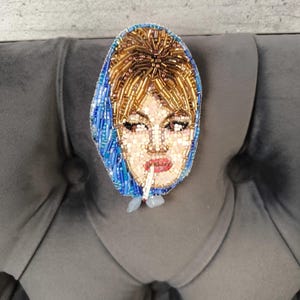 May include: A beaded brooch depicting a woman with blonde hair and a blue headscarf, smoking a cigarette. The brooch is made with small beads in shades of gold, blue, and pink, and is likely a handmade item. The brooch is a unique piece of jewelry.
