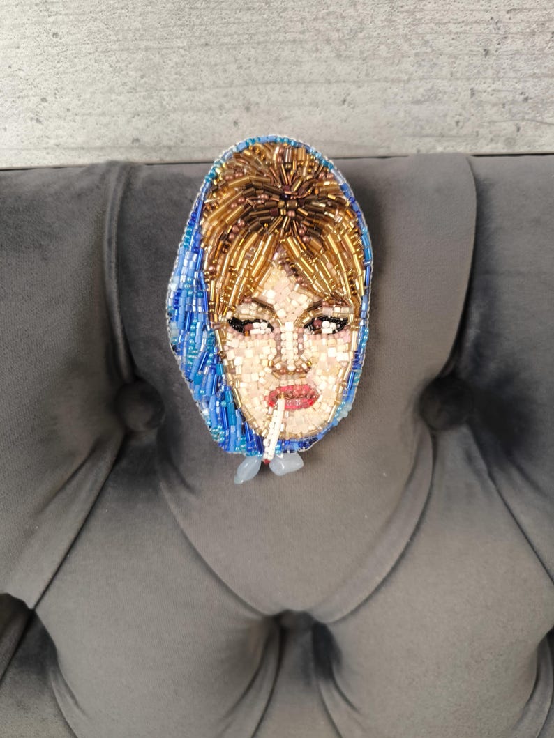 May include: A beaded brooch depicting a woman with blonde hair and a blue headscarf, smoking a cigarette. The brooch is made with small beads in shades of gold, blue, and pink, and is likely a handmade item. The brooch is a unique piece of jewelry.