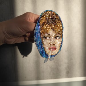 May include: A beaded brooch featuring a woman's face with a cigarette. The face is made of white, gold, and red beads, with blue beads outlining the headscarf. The brooch is held in a hand against a neutral background.