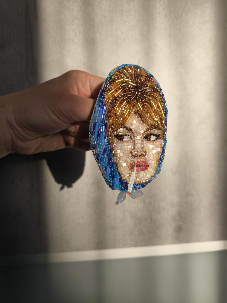 May include: A beaded brooch featuring a woman's face with a cigarette. The face is made of white, gold, and red beads, with blue beads outlining the headscarf. The brooch is held in a hand against a neutral background.