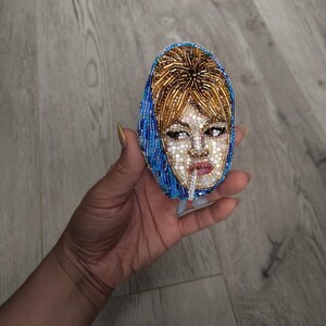 May include: Oval-shaped beaded brooch featuring a woman's face with a cigarette. The face is made of small white beads, with brown hair and a blue headscarf. The brooch is held in a hand.
