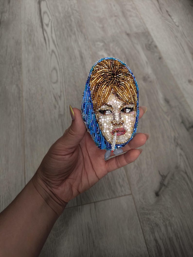 May include: Oval-shaped beaded brooch featuring a woman's face with a cigarette. The face is made of small white beads, with brown hair and a blue headscarf. The brooch is held in a hand.