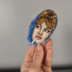May include: A beaded brooch depicting a woman with a cigarette. The brooch features a blue headscarf, blonde hair, and a detailed face. The brooch is made with small beads in various colors, creating a textured effect.