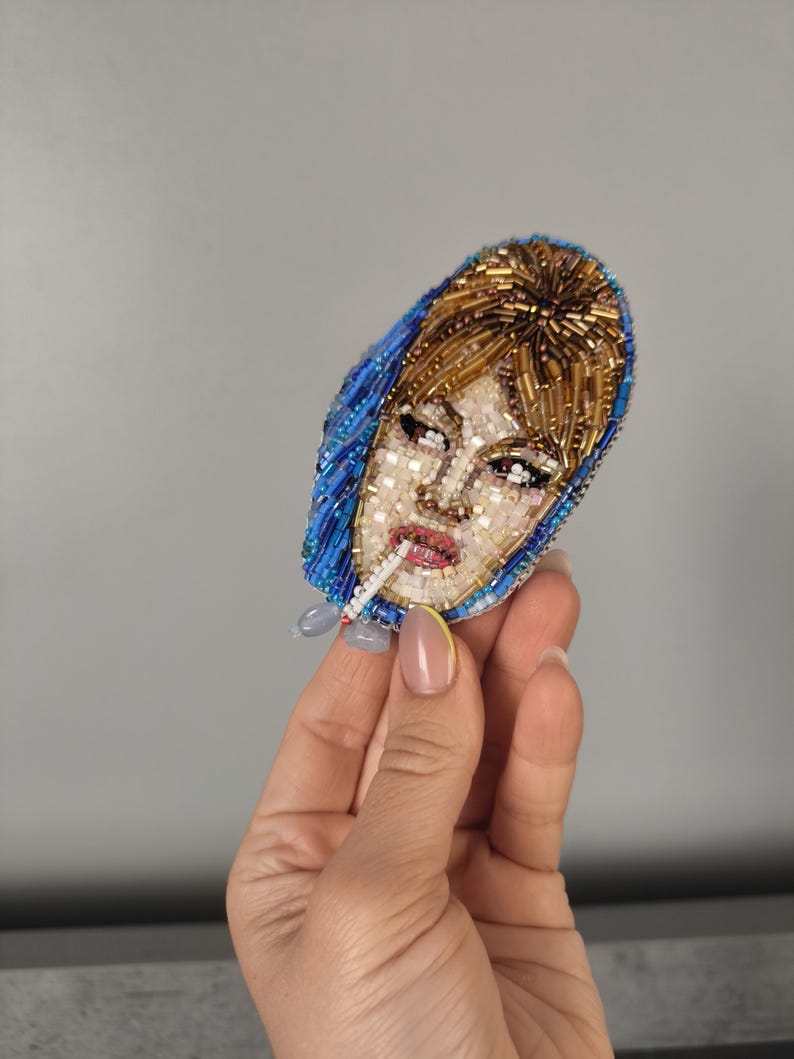 May include: A beaded brooch depicting a woman with a cigarette. The brooch features a blue headscarf, blonde hair, and a detailed face. The brooch is made with small beads in various colors, creating a textured effect.