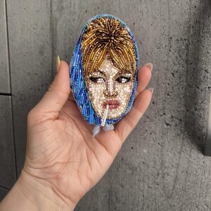 May include: Oval-shaped beaded brooch featuring a portrait of a woman with blonde hair and a blue headscarf. The brooch is made with small, colorful beads, creating a detailed image of the woman's face, including her eyes, lips, and hair. The brooch is held in a hand.
