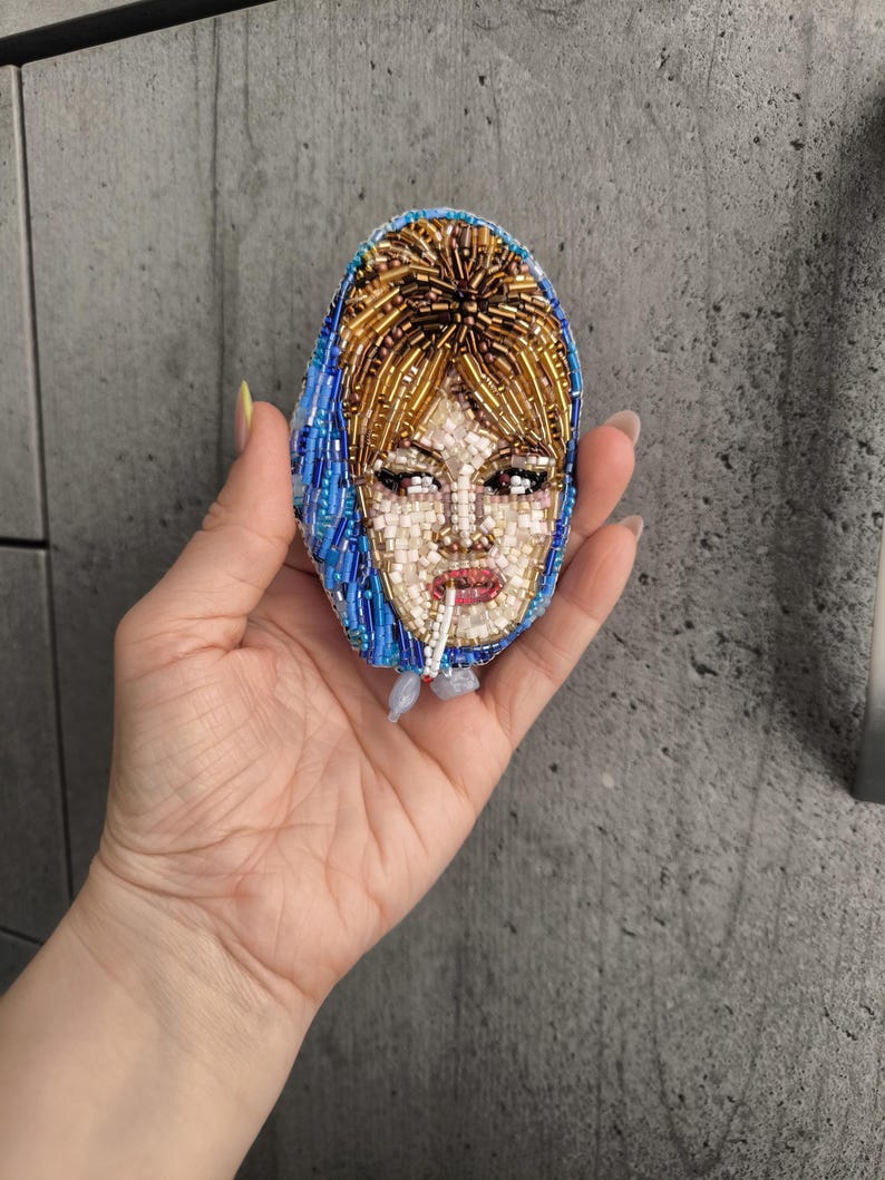 May include: Oval-shaped beaded brooch featuring a portrait of a woman with blonde hair and a blue headscarf. The brooch is made with small, colorful beads, creating a detailed image of the woman's face, including her eyes, lips, and hair. The brooch is held in a hand.