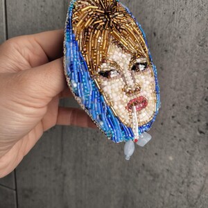 May include: A beaded brooch featuring a portrait of a woman with blonde hair and a blue headscarf, smoking a cigarette. The brooch is oval-shaped and made with various colored beads, including gold, white, blue, and red.