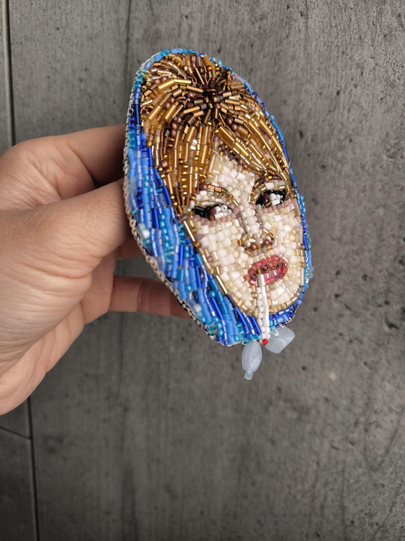 May include: A beaded brooch featuring a portrait of a woman with blonde hair and a blue headscarf, smoking a cigarette. The brooch is oval-shaped and made with various colored beads, including gold, white, blue, and red.