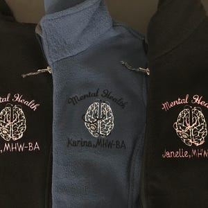 May include: Three fleece jackets in black and blue, each embroidered with "Mental Health" and a brain graphic. The black jackets have pink embroidery, while the blue jacket has white embroidery. Each jacket also has a name and credentials embroidered below the graphic.