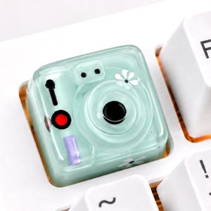 May include: A custom mechanical keyboard keycap shaped like a light mint green instant camera. The keycap features a black lens, a red shutter button, a purple flash indicator, and small white flower accents, positioned on a white keyboard.