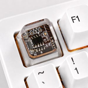 May include: A white mechanical keyboard featuring a custom transparent keycap that encapsulates a small brown circuit board with visible electronic components and printed text, positioned next to standard white keys labeled F1, F2, 1, 2, and Q.
