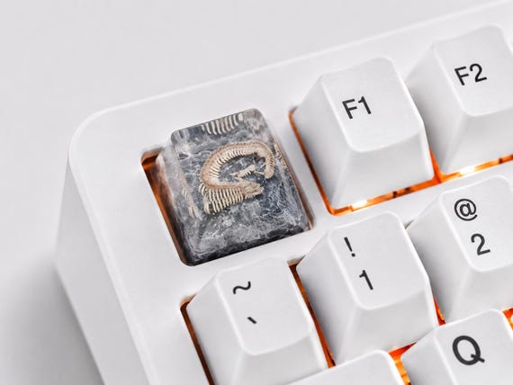 A hand-cast resin artisan keycap sculpted to look like a tiny gray bone fossil — the kind of hyper-niche desk flex that mechanical keyboard obsessives will lose their minds over.