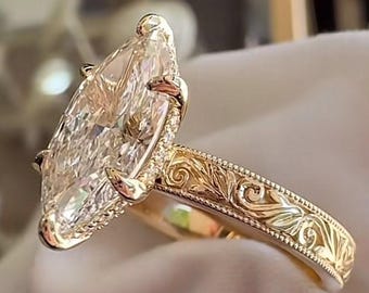 May include: A gold ring featuring a large marquise-cut center stone held by six prongs. The band is crafted from yellow gold and displays intricate, raised scrollwork engravings along the shank, bordered by fine milgrain detailing throughout.
