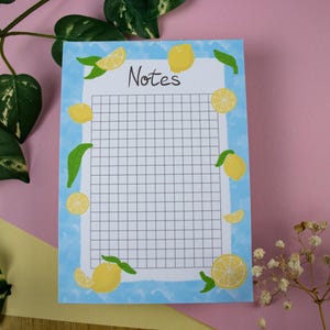 Könnte beinhalten: Ein rechteckiger Notizblock mit hellblauem Rand, verziert mit Illustrationen von Zitronen und grünen Blättern. Der Notizblock hat ein Rastermuster und das Wort "Notes" oben. Der Hintergrund ist eine Mischung aus Rosa und Gelb.