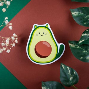 Könnte beinhalten: Ein skurriler Aufkleber mit einer katzenförmigen Avocado. Die Avocado ist hellgrün mit einem dunkelroten Kern und einem weißen Umriss. Der Aufkleber befindet sich auf einem weinroten und grünen Hintergrund mit Zweigen weißer Blüten und grünen Blättern.