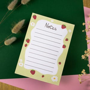 Könnte beinhalten: Ein hellgrüner Notizblock mit dem Wort "Notizen" in schwarzer Schrift. Der Notizblock ist mit roten Erdbeeren und weißen Blumen verziert. Der Notizblock befindet sich auf einem grünen und rosa Hintergrund.