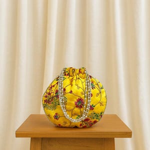 May include: A yellow, round drawstring bag with floral embroidery in red, green, and pink. A beaded strap hangs over the bag. The bag sits on a small wooden table against a cream-colored backdrop.