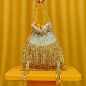 Puede incluir: Un bolso con cordón dorado y plateado con cuentas, un asa dorada y borlas. El bolso está sobre una superficie amarilla, sostenido por dos manos. El fondo es amarillo liso.