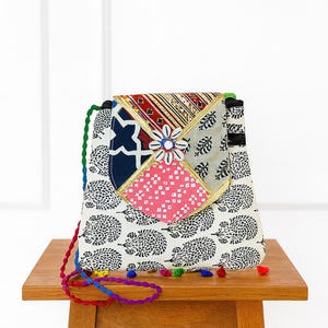 May include: A patchwork crossbody bag with a white background and black hedgehog pattern. The flap features a navy blue, red, and pink design with a flower embellishment. The strap is a colorful braided cord with tassels.
