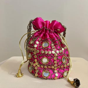 Könnte beinhalten: Ein leuchtend pinkfarbener Beutel mit Kordelzug, verziert mit goldenen Verzierungen und kleinen Spiegelakzenten. Der Beutel hat eine satinartige Textur und einen goldfarbenen, perlenbesetzten Kordelzug.