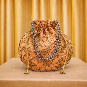 Puede incluir: Bolsa redonda color melocotón con bordados dorados y detalles de cuentas. La bolsa tiene una correa de cuentas y borlas doradas. El fondo es de un color dorado suave, lo que sugiere una estética lujosa.