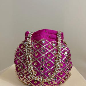 May include: A fuchsia-colored, round handbag with a diamond pattern, gold trim, and small mirrors. The bag has a pearl-like beaded strap. The bag is likely for special occasions.