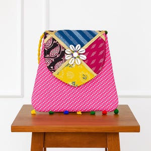 May include: A pink, patterned handbag with a patchwork design. The flap features a flower made of seashells. The bag has a yellow cord strap and is decorated with colorful pom-poms.