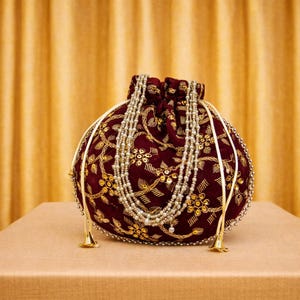 Może przedstawiać: Bordowa aksamitna torebka ze złotym haftem kwiatowym i sznurkiem ze sznurkiem z koralików. Torebka jest ozdobiona perłowym naszyjnikiem i złotymi frędzlami, tworząc elegancki dodatek.