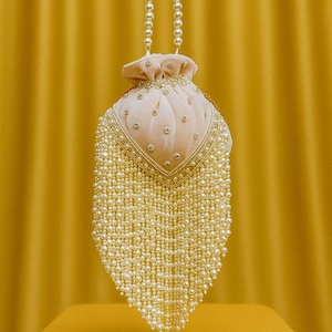 Puede incluir: Un bolso con cordón rosa rubor con flecos de perlas. El bolso presenta un asa con cuentas de perlas y está adornado con pequeños acentos brillantes. El bolso está sobre un fondo amarillo.