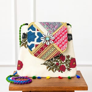 May include: Cream-colored handbag with a patchwork flap featuring geometric and floral patterns in red, blue, and gold. The bag has a green strap and is adorned with a seashell and turquoise embellishment. A colorful beaded bracelet and pom-poms are on a wooden stool.